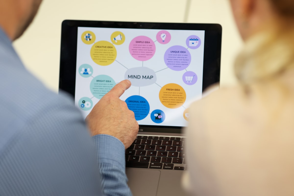 Two people viewing a mind map displayed on a laptop screen, one person pointing at the central idea labeled "MIND MAP," surrounded by colored circles containing various keywords related to creative thinking.